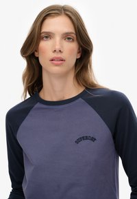 Jonge vrouw met lichtbruin haar draagt een blauw shirt in twee tinten met het "Superdry" logo op de borst tegen een effen witte achtergrond.