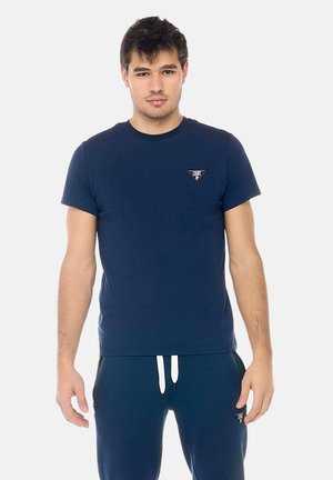 Giovane uomo in piedi di fronte, indossa una maglietta blu navy e pantaloni della tuta abbinati con cordini bianchi e piccoli loghi triangolari.