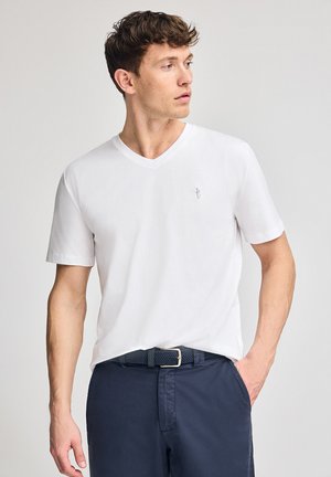 Camiseta de algodón blanca con cuello en V y mangas cortas, que presenta un pequeño logo bordado en el pecho, combinada con pantalones azul marino y un cinturón.