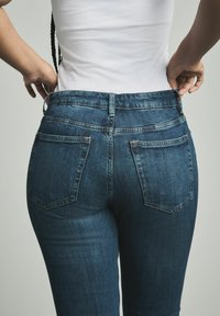Jeans en denim bleu foncé avec une coupe ajustée, cinq poches, coutures visibles et une taille mi-haute, vus de dos.