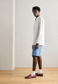 Hvitstripete skjorte med knapper, blå denimshorts, brune loafers og hvite sokker. Modellen står på lys tregulv mot en ensfarget vegg.