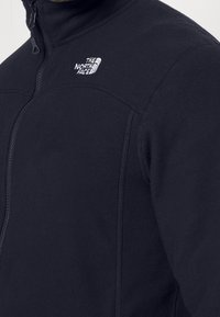 Giacca in pile nera con colletto alto, apertura con zip e logo bianco sulla parte superiore sinistra del petto. Caratterizzata da una texture liscia e un design aderente.