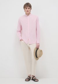 Helles rosa Langarmhemd mit Kragen, kombiniert mit cremefarbener Hose und schwarzen Sandalen. Ein Strohhut wird in der linken Hand gehalten.