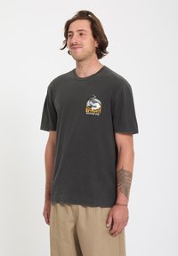 Homme portant un t-shirt Volcom gris foncé avec logo et un pantalon beige, debout devant un fond blanc uni, avec un tatouage de fougère sur l'avant-bras gauche.