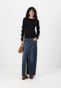 Schwarz langärmliges Oberteil mit Schleifenakzenten an den Ärmeln, kombiniert mit einem hochtaillierten Denim-Rock mit Schlitzen vorne. Die Accessoires umfassen eine braune Handtasche.