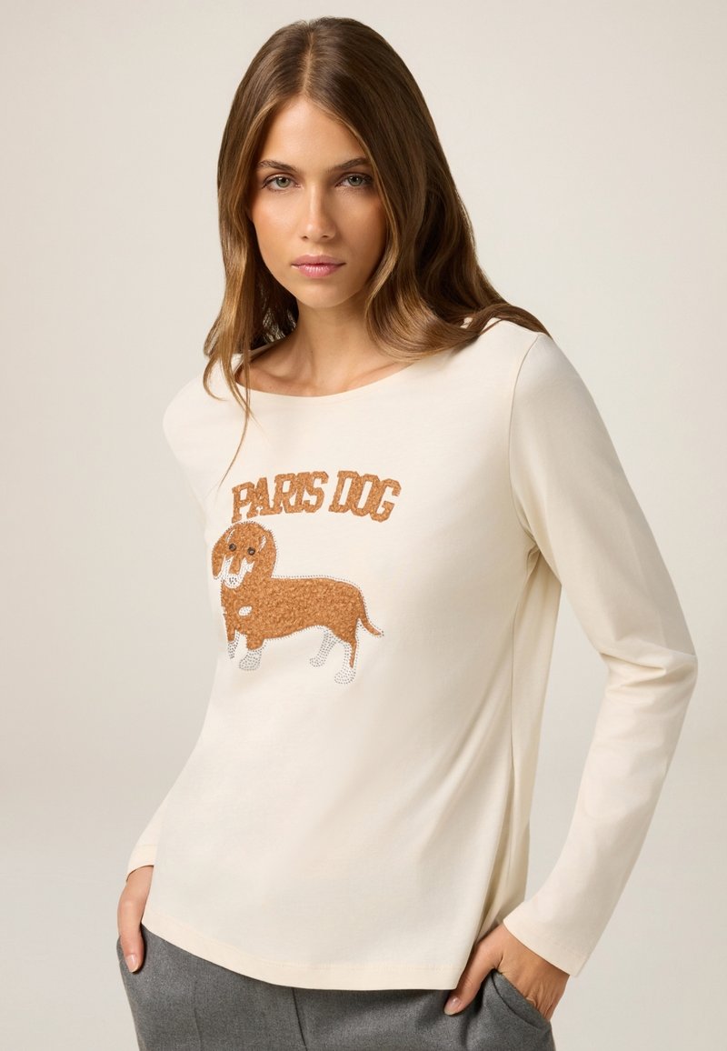 Camisa de manga larga color crema hecha de una tela suave, que presenta un dachshund bordado con el texto "PARIS DOG" encima en hilo marrón.
