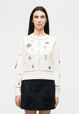 Žena s rovnými tmavými vlasmi nosí biely zapínaný sveter sčervenou lemovkou a viacfarebnými vyšívanými nášivkami, v kombinácii s čiernou sukňou s kvetinovými detailami.