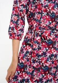 Vestido de patrones florales en tonos de rojo, rosa y azul. Presenta una cintura ceñida con un cinturón de tela atado y mangas de tres cuartos. Material texturizado.