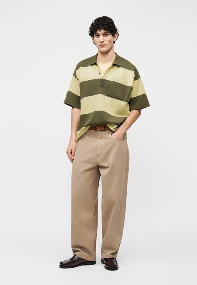 Νέος άνδρας που φοράει υπερμεγέθη ριγέ πόλο μπλούζα σε πράσινο και μπεζ, μπεζ παντελόνια με φαρδιά μπατζάκια, καφέ ζώνη και σκούρα δερμάτινα παπούτσια, στέκεται μπροστά σε απλό φόντο.