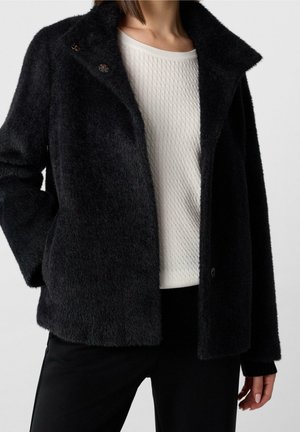 Femme portant un manteau noir duveteux sur un pull blanc texturé et un pantalon noir, debout avec une main dans la poche devant un fond uni.