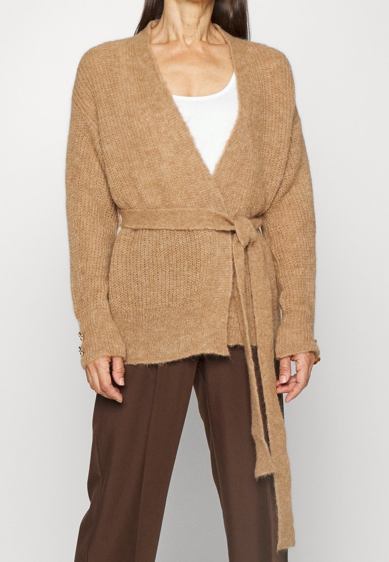 Cárdigan de punto beige con un diseño envolvente, mangas largas y un cinturón atado en la cintura. Presenta un acabado texturizado y sutiles detalles de botones.