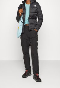 Svart isolerad jacka med huva, ljusblå skjorta, svarta cargo-byxor och svarta vandringsskor. Tillbehör inkluderar en ljusblå väska.