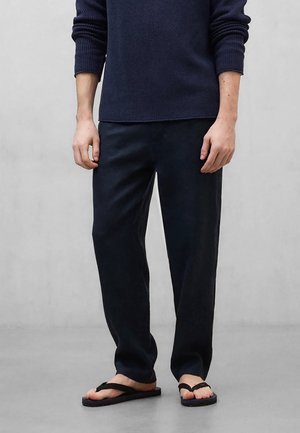 Mand iført navyblå sweater, løse mørke bukser og sorte flip-flops, stående mod en ensartet lys grå baggrund.