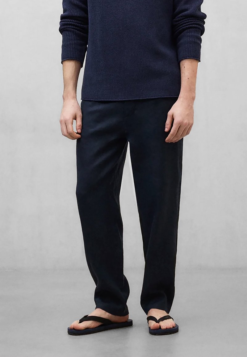 Homme portant un pull marin, un pantalon foncé ample et des tongs noires, debout devant un fond gris clair uni.