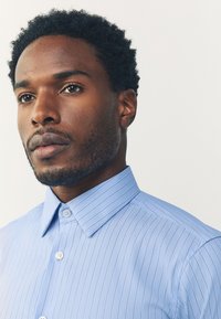 Camicia a righe azzurro chiaro con colletto a punta, caratterizzata da una texture liscia e piccoli bottoni lungo la patta frontale.