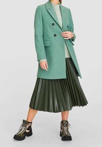 Cappotto doppiopetto verde in misto lana con rever sagomati, abbinato a una gonna plissettata in ecopelle verde scuro e stivali neri robusti.