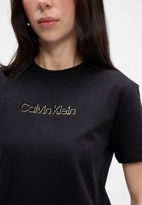 Čierne bavlnené tričko s krátkymi rukávmi, s vyšitým zlatým logom "Calvin Klein" na prednej strane, okrúhly výstrih, hladká textúra.