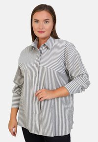 Chemise à rayures grises et blanches avec un col pointu, des manches longues et un détail de poche avant. Texture en tissu doux et tissé.