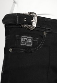 Crne traperice od denima s oznakom Versace Jeans Couture, s teksturiranim srebrnim ukrasnim kopčanom za pojas i kontrastnim detaljima šavova.