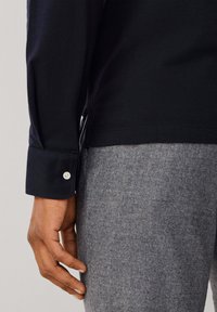 Mornarska košulja s dugim rukavima, dugmadi na manžetama i bočnim prorezima, u kombinaciji s grimiznim hlačama teksturiranog izgleda, koje prikazuju glatku završnu obradu tkanine.