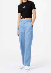 Woman wearing a black cropped T-shirt with a logo, light blue high-waisted wide-leg pants, and white sneakers, standing with one hand in pocket.