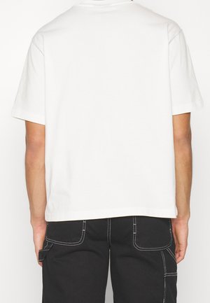 Biała bawełniana koszulka, krótki rękaw, okrągły dekolt, luźny krój. Widok z tyłu pokazuje gładki fason, bez wzorów czy akcentów, zestawiona z czarnymi spodniami.