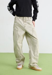 Lys beige baggy jeans med en falmet tekstur, har store sidelommer og en knaplukning, kombinert med sneakers og en svart langermet skjorte.