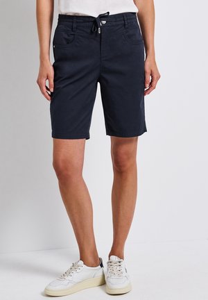Vrouw draagt marineblauwe knielange shorts met voorzakken en een tailleband met trekkoord, gecombineerd met witte sneakers en een wit bovenstuk.
