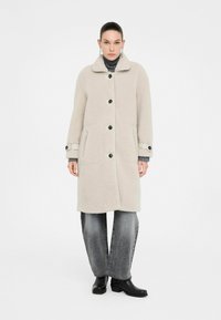 Donna che indossa un cappotto di lana beige con bottoni, un maglione a collo alto grigio, jeans larghi neri sbiaditi, stivaletti neri e orecchini a goccia.