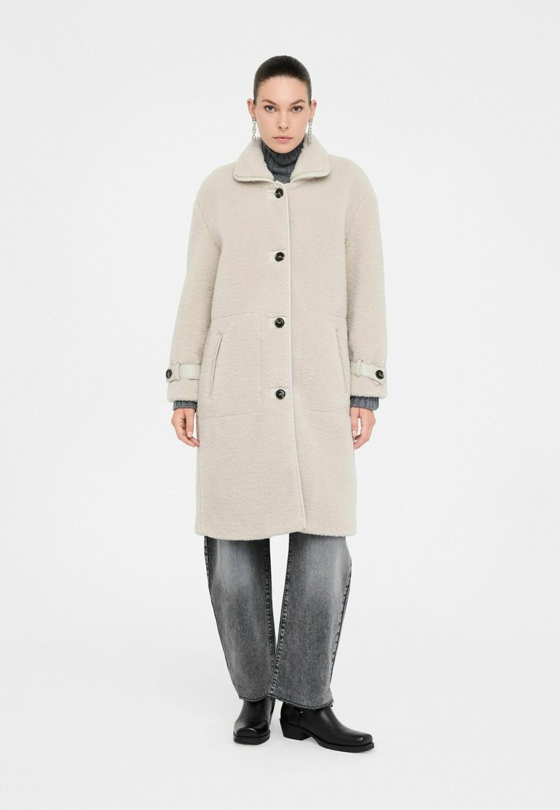 Donna che indossa un cappotto di lana beige con bottoni, un maglione a collo alto grigio, jeans larghi neri sbiaditi, stivaletti neri e orecchini a goccia.