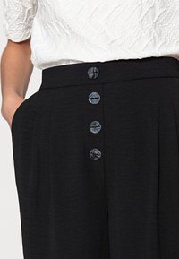 Zwarte broek met een hoge taille, gestructureerde stof en vijf decoratieve knopen aan de voorkant. Zakken aan de zijkant zichtbaar.