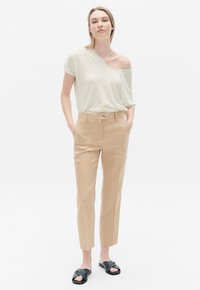 Top grigio chiaro monospalla abbinato a pantaloni beige sartoriali. I pantaloni presentano una chiusura con bottone e tasche laterali. Scarpe nere senza lacci.
