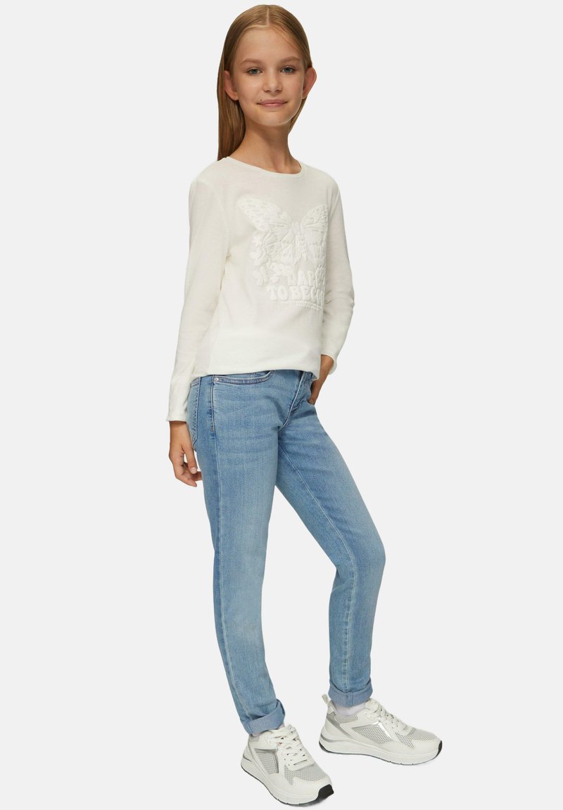 Witte blouse met lange mouwen, voorzien van een vlinder- en tekstontwerp, gecombineerd met lichtblauwe skinny jeans en witte sneakers. Zachte textuur over het geheel.