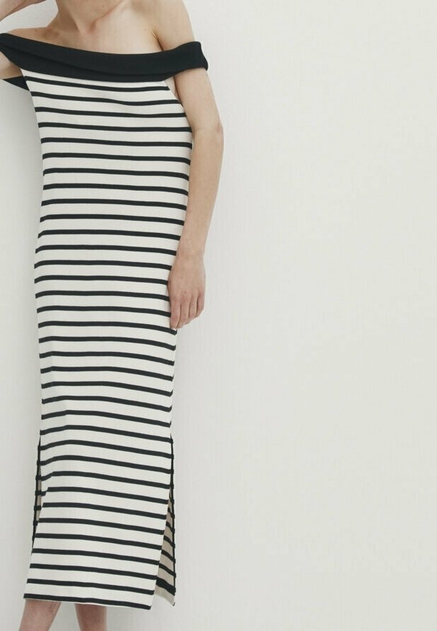 Femme portant une robe maxi à rayures horizontales noires et blanches, épaules dénudées, avec des fentes latérales, se tenant devant un fond clair uni.