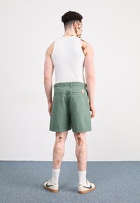 Pantalones cortos verdes de corte relajado, con bolsillos traseros, combinados con una camiseta sin mangas blanca y zapatillas Nike blancas. Fondo liso y moqueta azul.