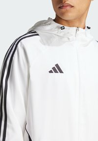Veste blanche légère avec capuche, ornée de rayures noires le long des épaules, d'une fermeture éclair et d'un logo noir brodé sur la poitrine.