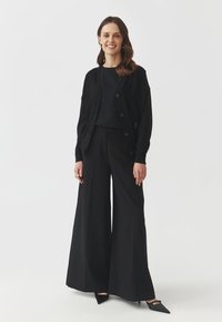 Černý outfit složený z k cardiganu na knoflíky, přiléhavého topu a širokých nohavic. Celkový vzhled doplňují lesklé lodičky s špičatou špičkou. Hladká tkanina, minimalistický design.