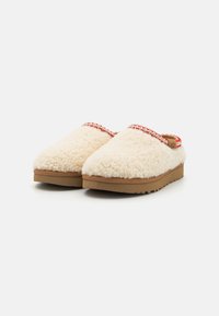 Beige fluffy klipklapper med en tekstureret pelsoverdel, rød fletkant og en robust brun gummisål for komfort og greb.