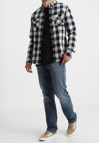 Camicia flanella a scacchi bianco e nero sopra una maglietta nera, abbinata a jeans blu e sneakers beige. Due tasche frontali visibili.