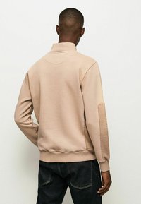 Sudadera beige con cuello alto, puños acanalados y textura suave. Presenta un panel texturizado en la manga izquierda. Llevada con jeans oscuros.