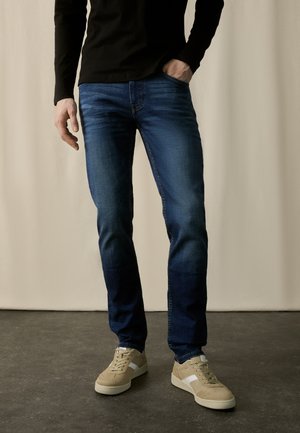 Dark blue jeans with a slim fit, featuring a slight fade and five-pocket design, paired with beige sneakers with white accents.