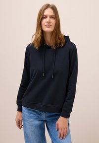 Kvinde iført en sort hættetrøje med snore og blå jeans, stående mod en ensfarvet beige baggrund.