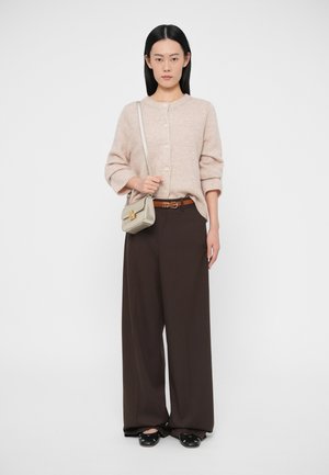 Žena stojící v béžovém propnutém svetru na knoflíky, hnědých širokých kalhotách, černých balerínách a s béžovou kabelkou přes rameno se zlatou sponou.