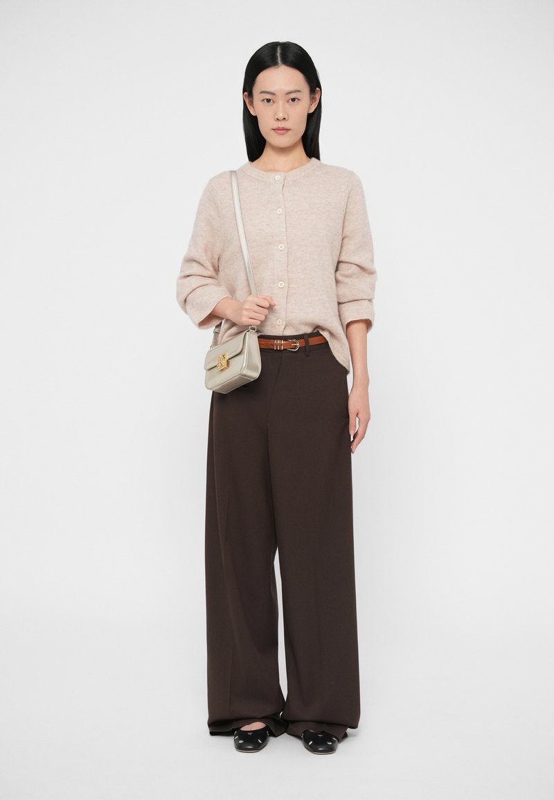 Ženska stoji oblečena v bež gumbasti pulover, rjave široke hlače, črne balerinke in bež torbico sramo zlato zaponko.