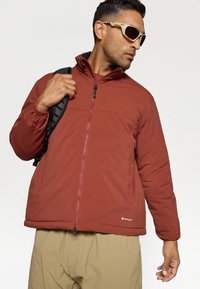 Veste isolante rouge avec une texture lisse, un col montant, une fermeture éclair à l'avant et des poches latérales. Assortie avec un pantalon kaki et des accessoires dans des tons neutres.