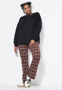 Sudadera negra de gran tamaño con mangas largas, combinada con pantalones de color burdeos con un patrón que presenta diseños en dorado y blanco. Zapatillas blancas con cordones negros.