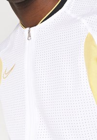 Nahaufnahme einer Person, die eine weiße Sportjacke mit Reißverschluss trägt, die kleine Perforationen aufweist, mit goldenen Nike-Logo und goldenen Akzenten an den Ärmeln.