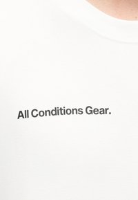 Άσπρο μπλουζάκι με στρογγυλή λαιμόκοψη και μαύρο κείμενο «All Conditions Gear.» στο κέντρο του στήθους.