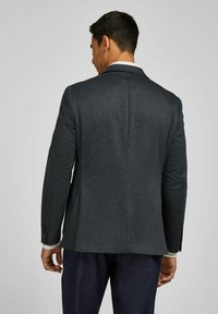 Mann mit dunkel strukturiertem Blazer und weißem Hemd, der von einem schlichten hellen Hintergrund abgewandt ist.
