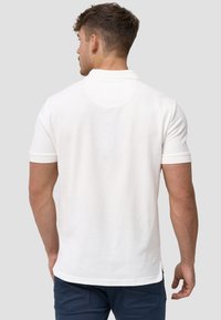 Weiße Poloshirt aus strukturiertem Stoff, mit kurzen Ärmeln und flachem Kragen. Die Rückansicht zeigt eine glatte Verarbeitung und subtile Nähdetails.